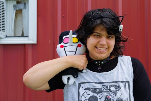 Youung woman holds a plushie with a long nose were her arm clamped around it with a dark shirt under a light grey shirt and a wry smile, medium length dark hair with a transparent cat ear headband; behind a red metal barn wall and i think a window with an air condition