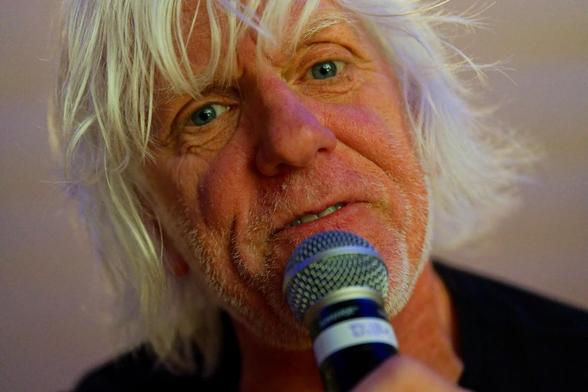 Man with white hair holding microphone makes eye contact at close range and fills most of the frame