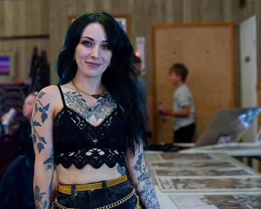 Woman with dark hair and Dark lacy top showing leaf-like tattos with necklace and open midriff and thin beige belt holding up a denim skirt or pants encircled by a studded band with rock and roll posters laid out on a table to her right and a blurred out boy walking by a door and a vertically striped wall