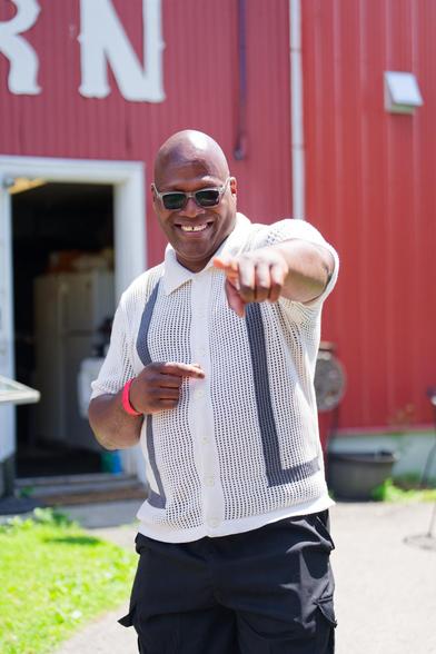 Black man white with white shirt with dark stripes and dark pants points at me and smiles wearing dark sunglasses in front of a red barn sort of a building