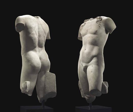 Photograph of a Roman marble torso, circa 1st century BCE-1st century CE. Depicted lifesized, standing in contrapposto with his weight on his right leg, the left slightly advanced, with both arms lowered, the left pulled slightly back, the lithe figure with softly-modelled musculature, his long serpentine tendrils falling onto each shoulder, preserving a tree trunk support along the right thigh, and a smaller rectangular strut at the back of the left thigh. The position of the arms suggests tha…