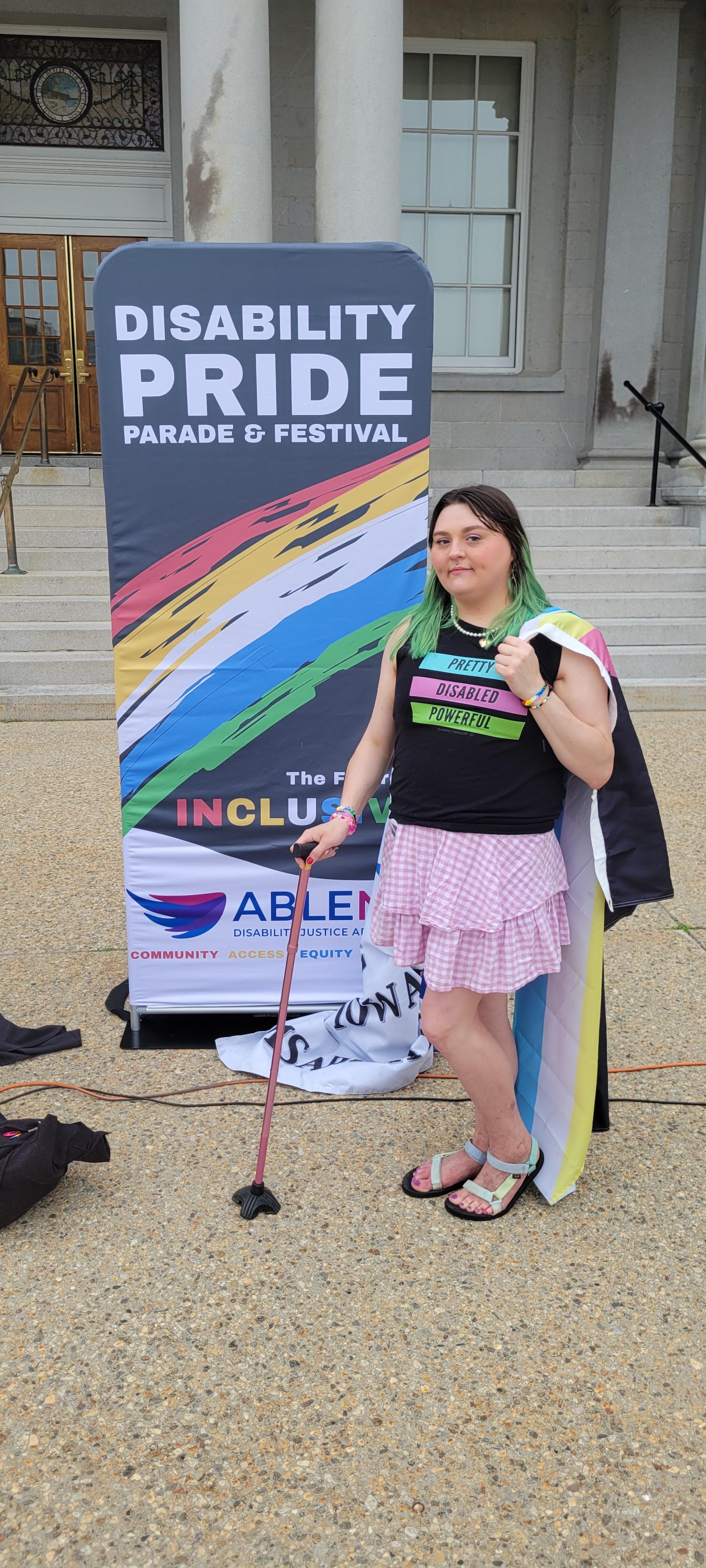 Picture of me at 2026 disability pride
 I'm standing outside in front of a poster for the event, I'm wearing a shirt that says "pretty disabled powerful".  I have my pink cane, with the disability pride flag thrown over my shoulder. I am looking at the camera smiling. My hair is green