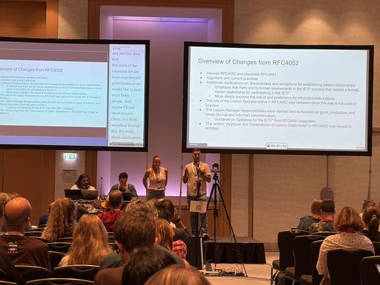 The image shows a presentation at a conference, with a speaker addressing the audience. Two projectors display slides summarizing changes to RFC4052 and RFC4691, focusing on clarifications for liaison relationships and responsibilities within the IETF process.