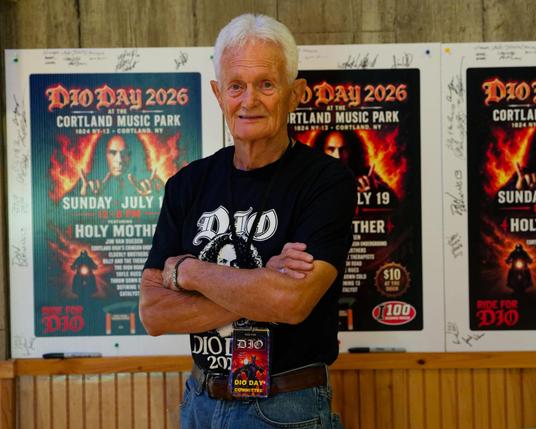 Man with white hair and crossed arms is wearing a black shirt that says DIO and has a picture of Runnie James Dio and there are three posters behind him with pictures of Dio too.