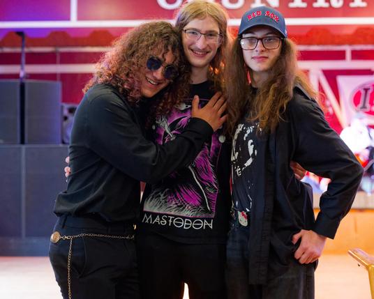 Three young men with long hair and dark clothes pose in front of stage