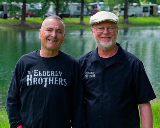 Two men with white hair and beards wear dary shirts that say the Elderly Brothers with a small pond behind and campers behind even that