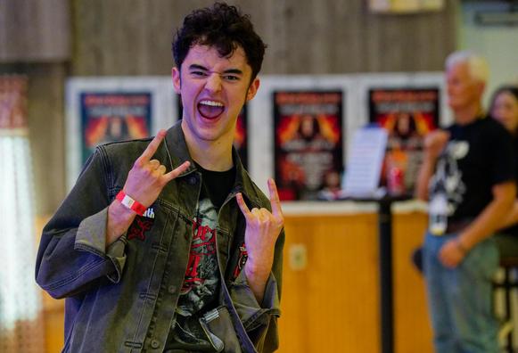 Man poses like he is screaming making the sign of the horns with both hands, a row of four dio posters is behind him and one of the organizers of the event looks on