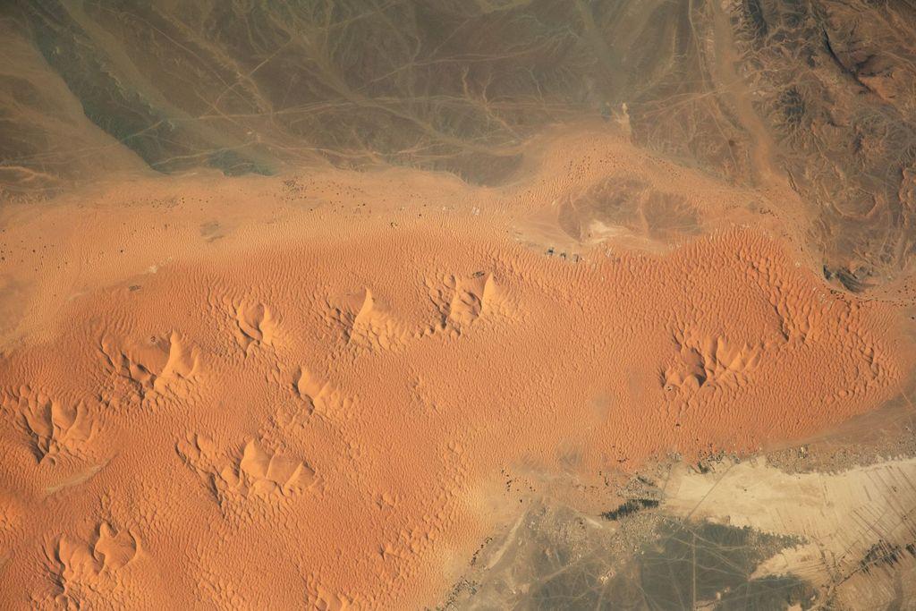 Vast aerial view of an orange sand sea of crescent and star dunes meeting a darker rocky plateau, highlighting wind-sculpted ripples, sharp dune crests, and the sandy margin dotted with small settlements and vehicle tracks.