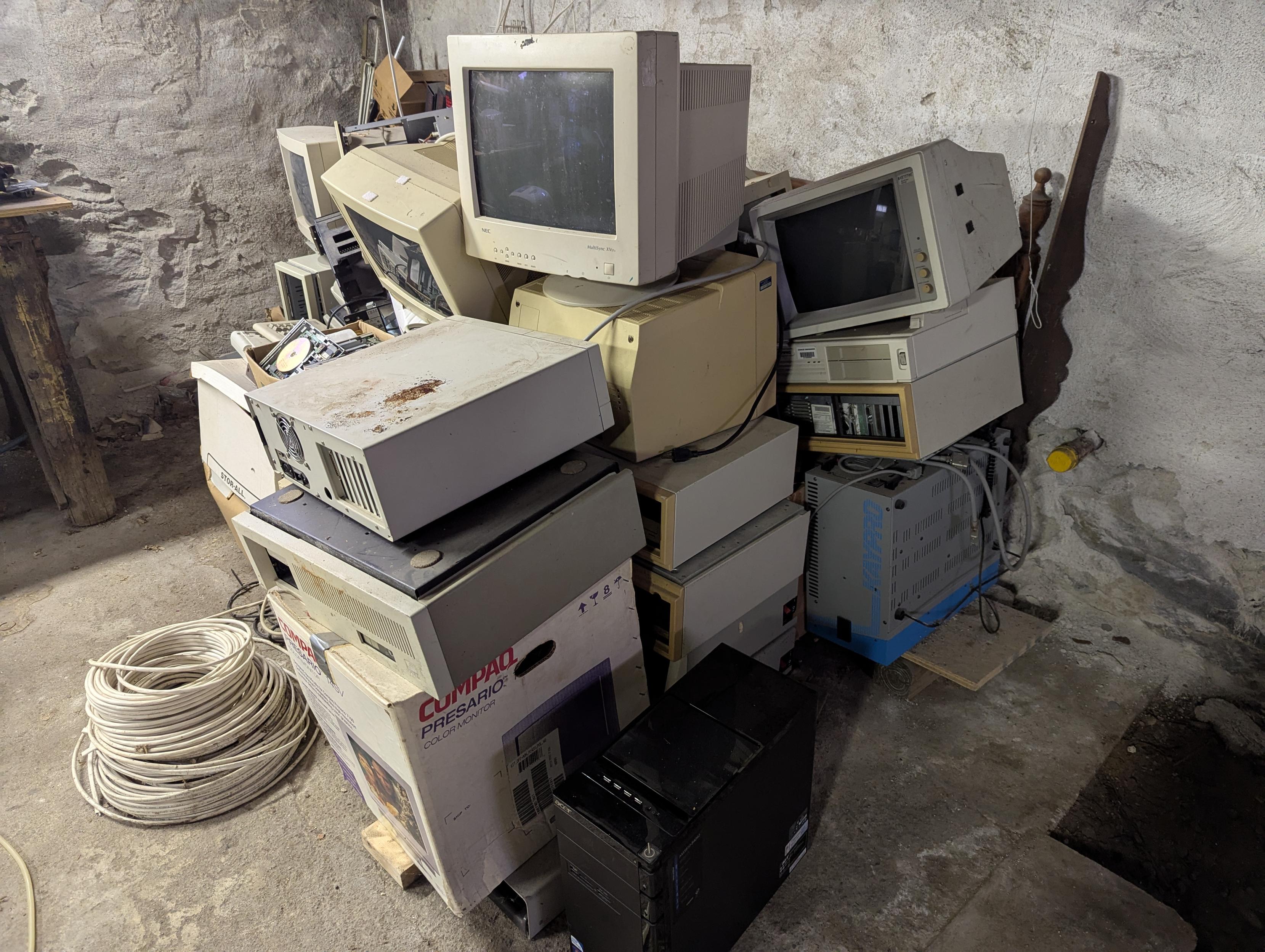 A literal pile of computers and monitors from the '80s and '90s, on a rough concrete floor in a cellar.