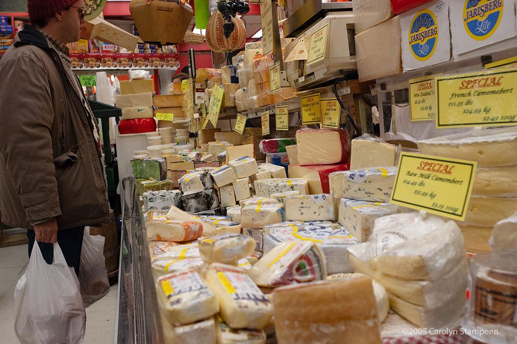 A man is holding two full plastic shopping bags and is looking over a huge selection of cheeses, many wheels or parts of wheels.