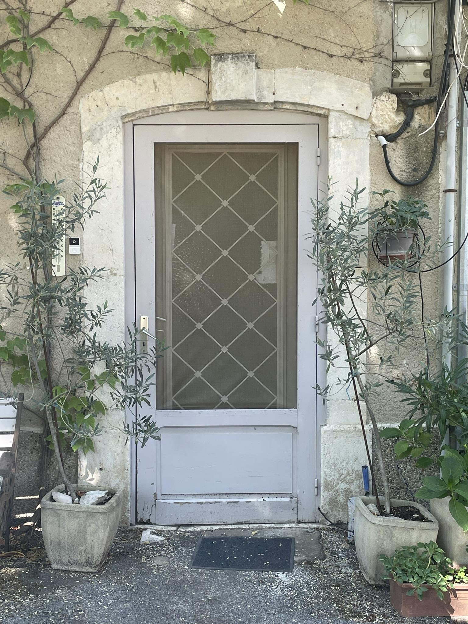 Porte vitrée avec croisillons en métal et moustiquaire, quelques pots de plantes devant.