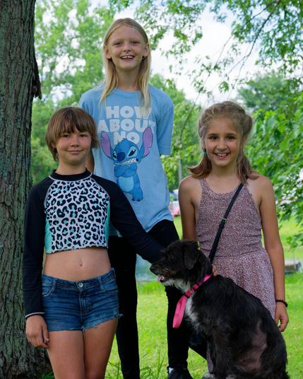 Three Children pose with a dog with lush green vegetation behind them.