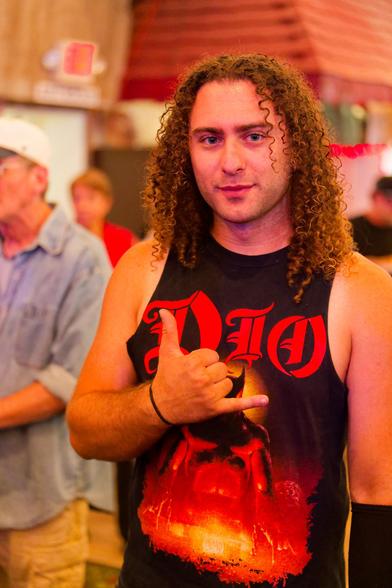 Man with very curly long hair wears a DIO tank top and makes the sign of the horns