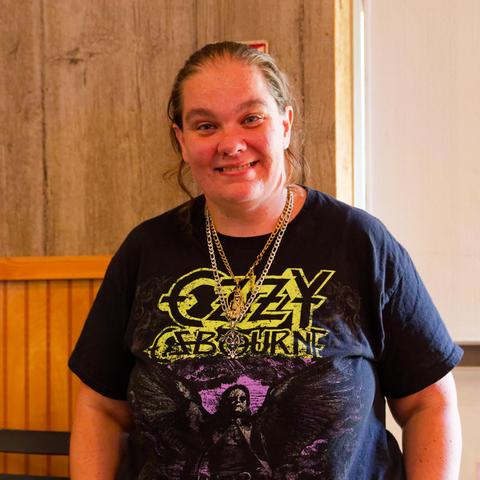 Smiling woman with at least two chain necklaces wears a black shirt that read Ozzy Osbourne in styling yellow text and then in purple and black below we see him wearing great big wings.