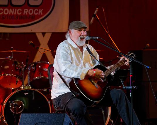 Man is sitting in front with a white shirt, red band to get in, grey cap, dark pants and a black-to-brown faded guitar with a drum set but no drummer in the background