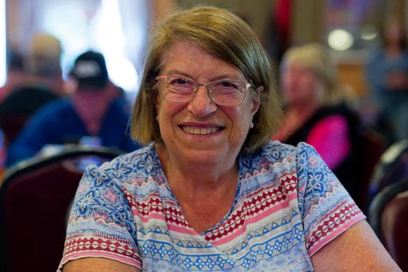 WOman wears a big smile, glasses, light hair in a bob haircut, and a short-sleever shirt with geometric red, blue and white patterns.