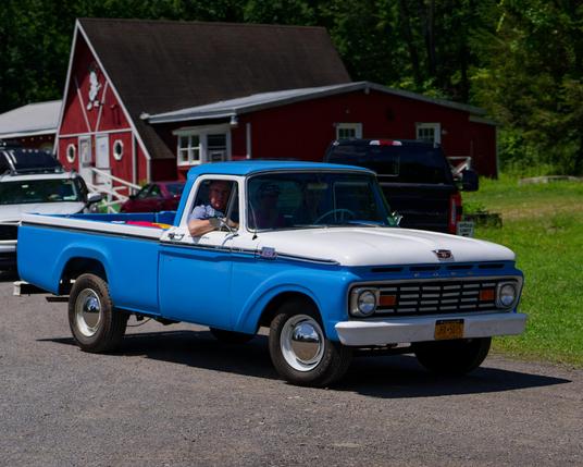 Old pickup truck drives in with mostly blue body with a white hood and accents and whitewall towers,  a red building with an A-frame center and two extensions on each side is behind it.