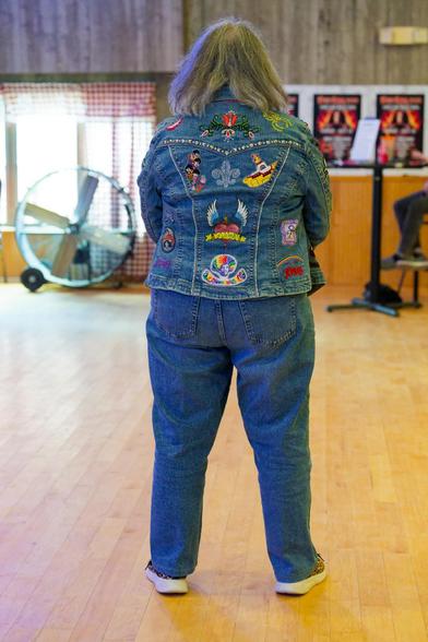 Woman with grey hair is standing away from me and showing off jacket with numerous patches from rock bands including the Beatl'es Yellow Submarine and Dio with a Ranibow.  She's standing on a wood dance floor with a window with a big fan in front and a row of dio day posters on the other side.