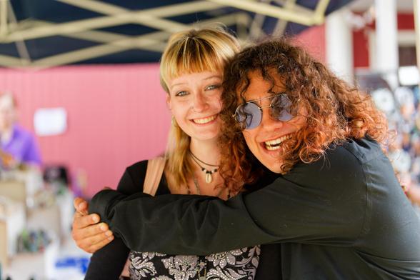 In the middle a blond girl wearing multiple necklaces, an open black jacket and a sparkly black and silver top and she's being hugged by a young man wearing a dark shirt and dark glasses and long thick curls.  Both are smiling,  the wall behind looks pink and above there are geometric structures on the ceiling.