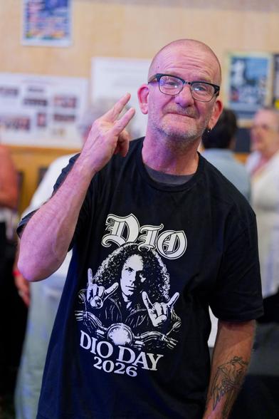 Older man with a tattoo on his arm makes sign of the horns while wearing a DIO DAY 2026 T-Shirt where Dio is making two of them,  some people and posters on the wall can be seen blurred out behind him.