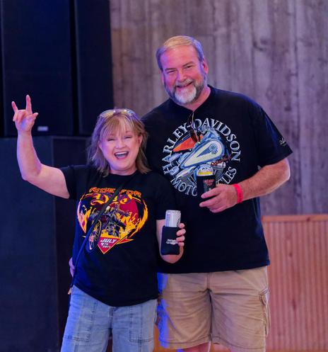 Woman with moderate length blond hair wearing black shirt with red-yellow heart-shaped motorcycle graphic makes sign of horns and holds beer;  to the right of her is a taller man also holding a beer with a Harley-Davidson T-shirt