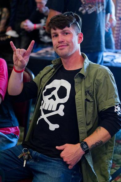 Man with short hair and sunglasses over his head makes sign of the horns with a green jacket with a skull and crossbones shirt underneath