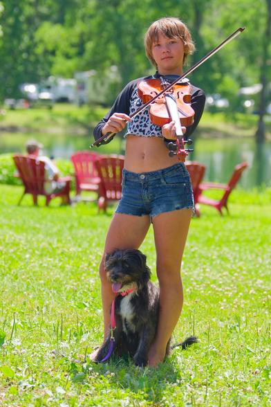 Girl with bob haircut holds violin and bow with a little dog between her legs on a clover-rich lawn with red chairs, a pond, and campers behind