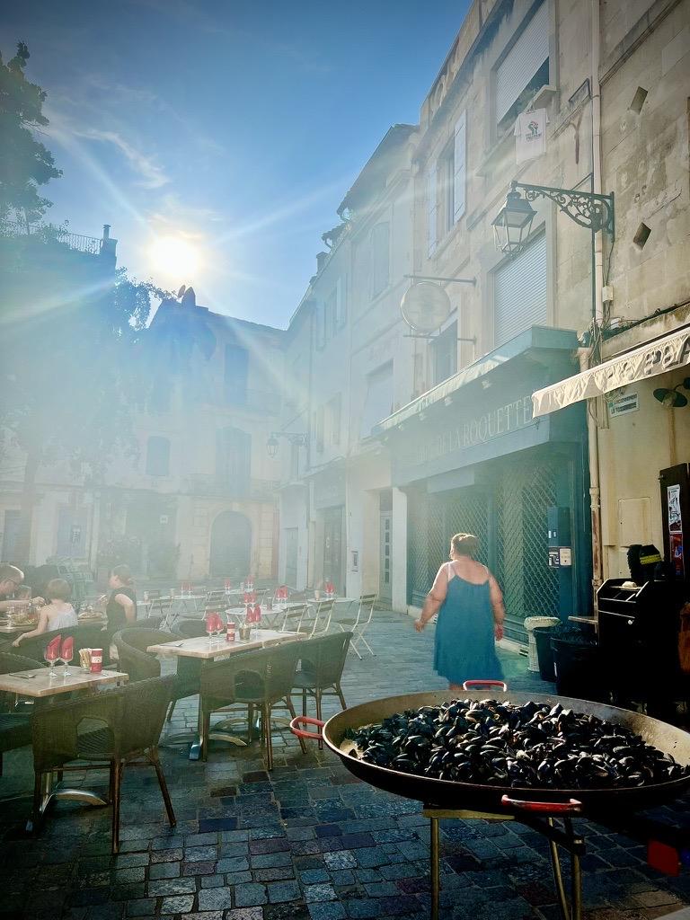 ‎⁨Arles⁩, ⁨Côte Camarguaise⁩, ⁨France⁩

A sun-drenched outdoor restaurant terrace features a massive, shallow metal pan filled with mussels in the lower right foreground. In the midground, a person in a teal dress walks past a store with a dark blue facade and an ornate street lamp. To the left, several empty wooden dining tables are set with red glasses, and a few people are seated at a table further back. Intense, hazy sunlight streams down from the upper left, casting a strong glare across the historic paved street and stone buildings.