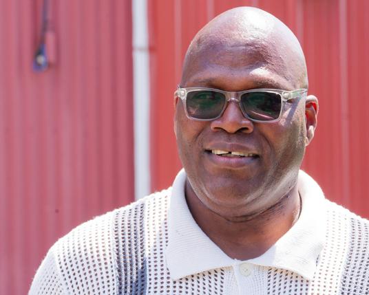 Black mand with shades stands in front of red barn with corrugated metal walls