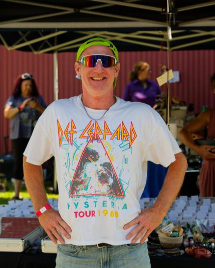 Man in wraparound shades with a flourescent green billed cap turned backwards arms akimbo wearing a white Def Leppard shirt promoting the Hysteria tour under the tent of a crystal vendor with corrugated red metal in the far background