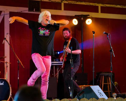Man in moderate length white hair sings into microphone wearing black MTV t-shirt with mushrooms and pink pants with a guitarist behind him