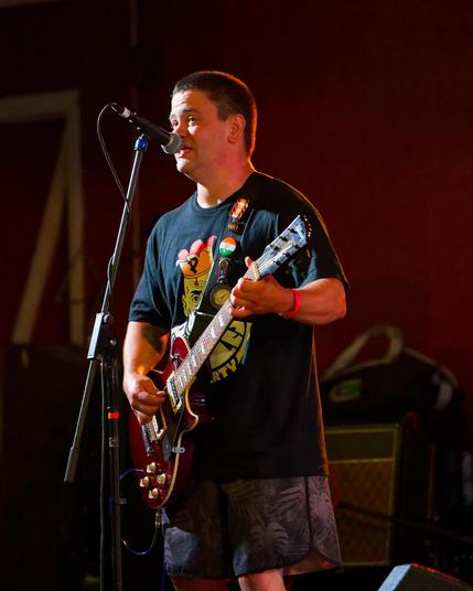 Man wearing black shirt with a yellow figure on it playing a six string guitar and playing into a microphone.