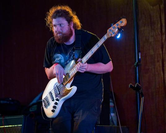 Man wirth reddish-brownish frizzy hair (backlit) with a thick beard wearing a black shirt and playing a white guitar