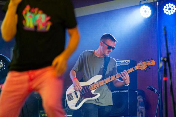 In the center a man in a medium grey shirt is playing a white bass guitar with lights behind him,  to the left in the front is a blurred out man with pink pants and an Mtv mushroom shirt.