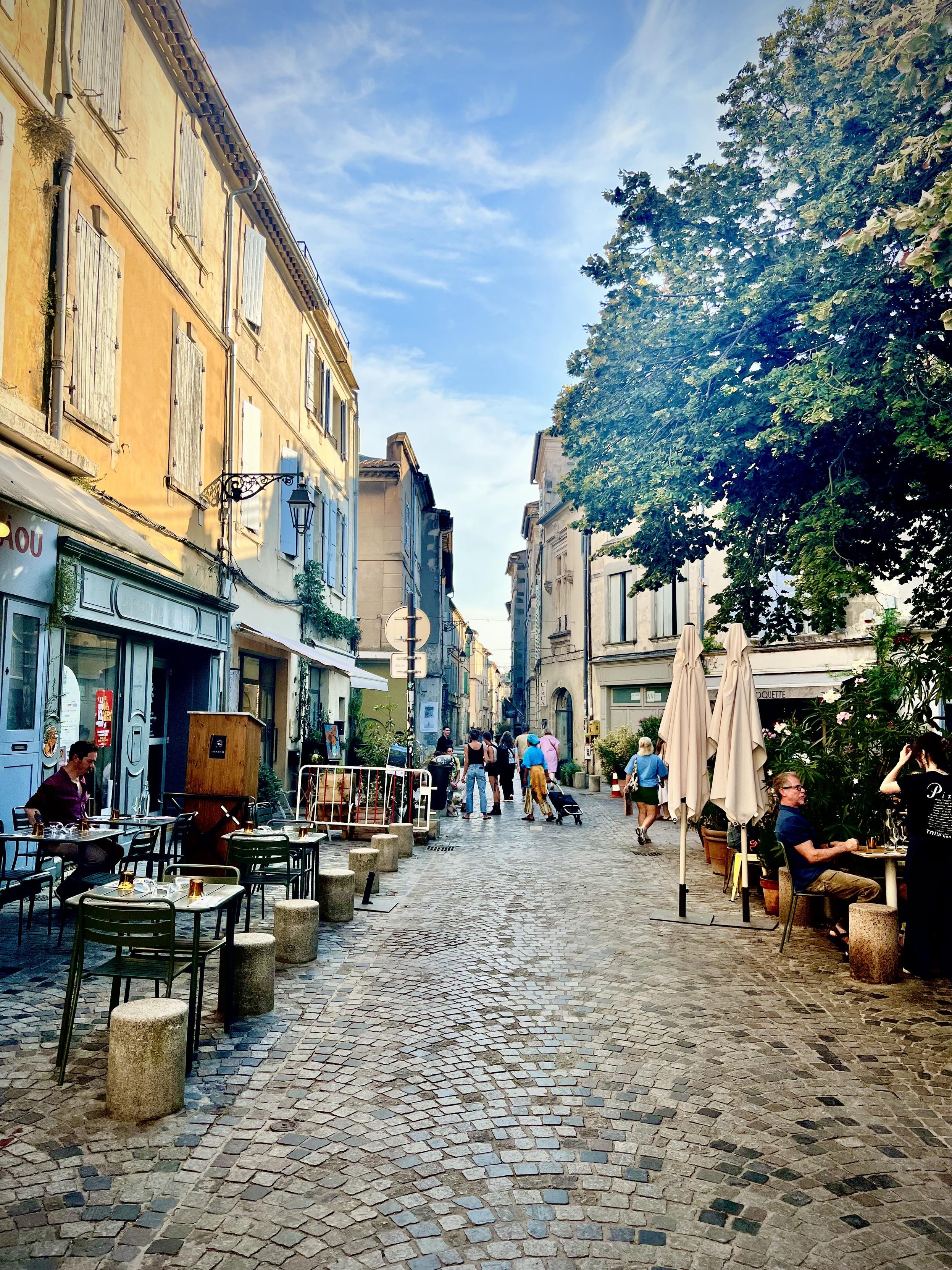 ‎⁨Arles⁩, ⁨Côte Camarguaise⁩, ⁨France⁩

A narrow cobblestone street stretches through a European town lined with historic plaster buildings featuring wooden shutters. Outdoor dining areas with tables, chairs, and closed umbrellas flank both sides of the pathway, where several people sit outside while pedestrians walk down the center of the street. Lush tree foliage partially frames the upper right under a bright blue sky with soft white clouds.