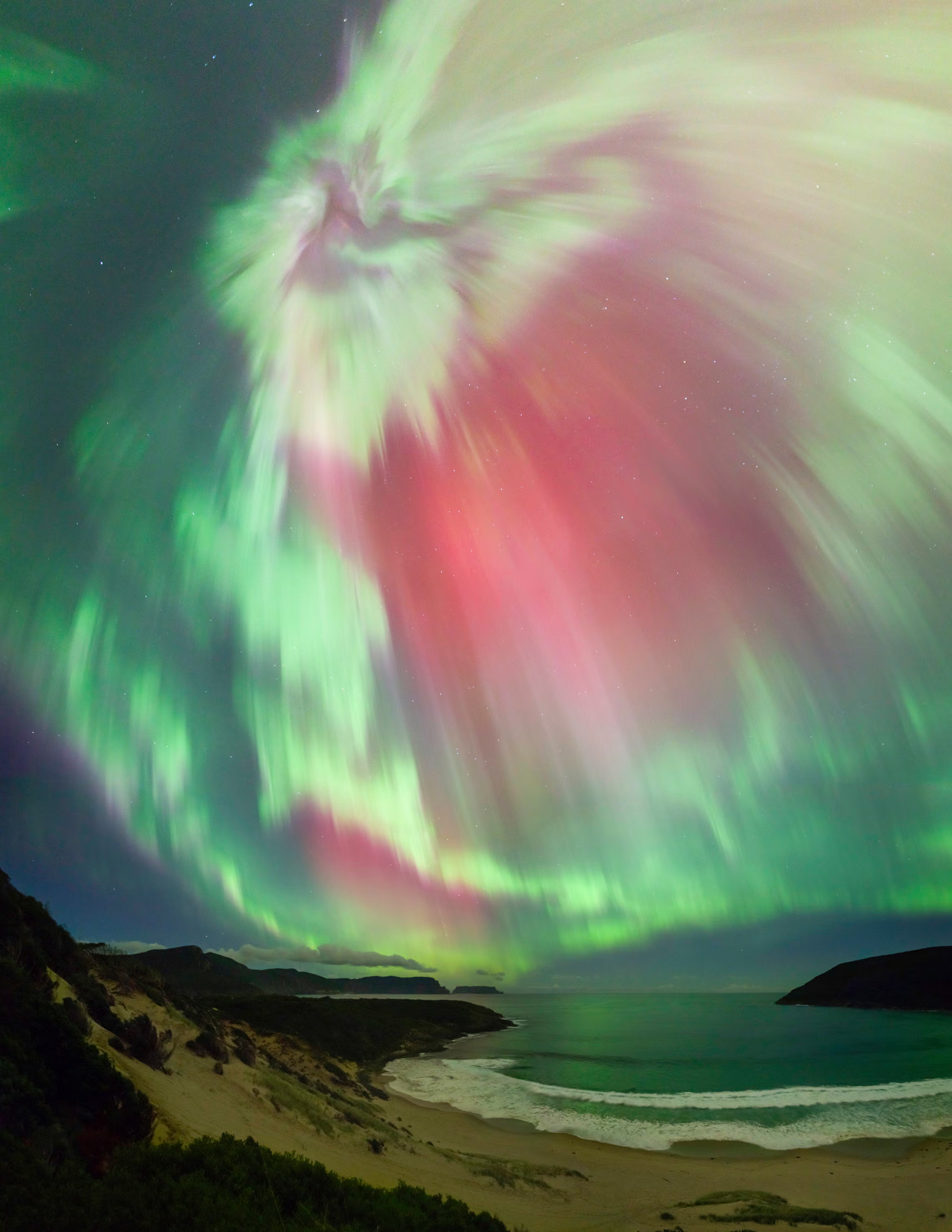 A vertical wide-angle photograph capturing a stunning aurora over a dark, sandy beach and bay at night. Brilliant white, magenta-pink, and emerald-green rays of light burst outward in a starburst pattern from a central point high in the night sky. The glowing green lights fan down toward a dark coastline where gentle waves roll onto a sandy shore. Dark, grassy hills frame the left and right sides of the beach under the illuminated sky.