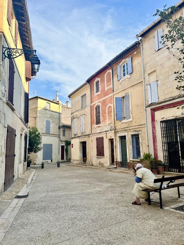 ‎⁨Arles⁩, ⁨Côte Camarguaise⁩, ⁨France⁩

A wide, cobblestone or gravel street curves between historic multi-story buildings with warm-toned plaster and stone facades, adorned with wooden shutters in blue, brown, and gray hues. On the right foreground, a person wearing light-colored traditional clothing and a white hat sits on a wooden bench looking down at a mobile device. Warm sunlight highlights upper portions of the buildings against a soft blue sky with light clouds.