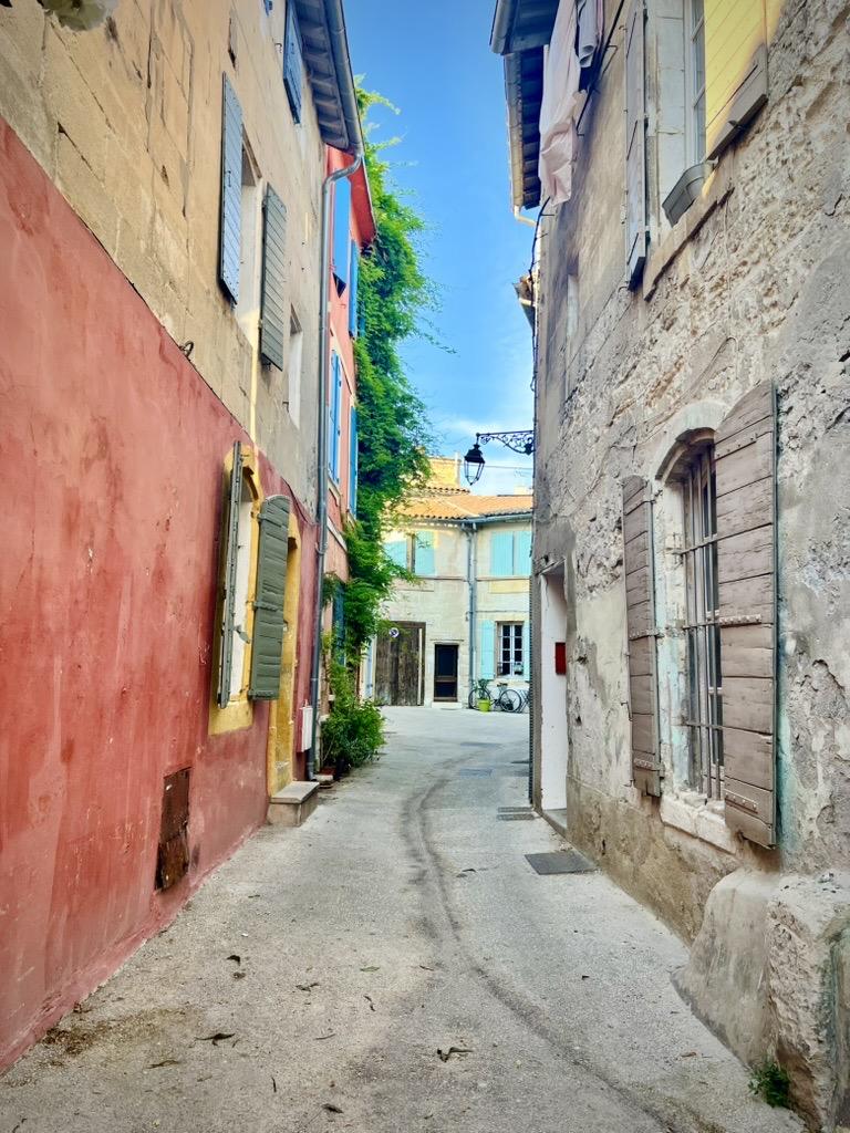 ‎⁨Arles⁩, ⁨Côte Camarguaise⁩, ⁨France⁩

A narrow street curves gently between old buildings, featuring a reddish-pink plastered wall on the left and a weathered stone facade with wooden shutters on the right. Green vines climb up a multi-story building frame in the mid-ground, leading toward an open courtyard in the distance under a bright blue sky.