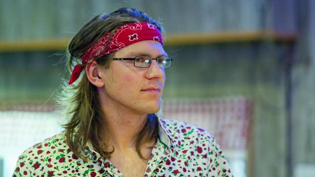 Man with hair just past shoulders and red bandanna, thin glasses and shirt with red roses with green leaves on white background with window with checkerboard curtains behin dhim