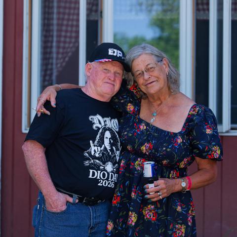Older man and woman:  on the left a man with dark cap that reads EJP and dark shirt that read DIO DAY 2026 and on the left a woman with grey hairand dark dress with colorful floral-like pattern