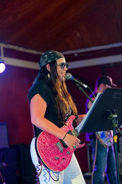 Woman with hair about halfway the length of her torso strums a pink tiger striped guittar and sings into a microphone:  she has a black bandanna, black shirt with very short sleeves and light denim pants.