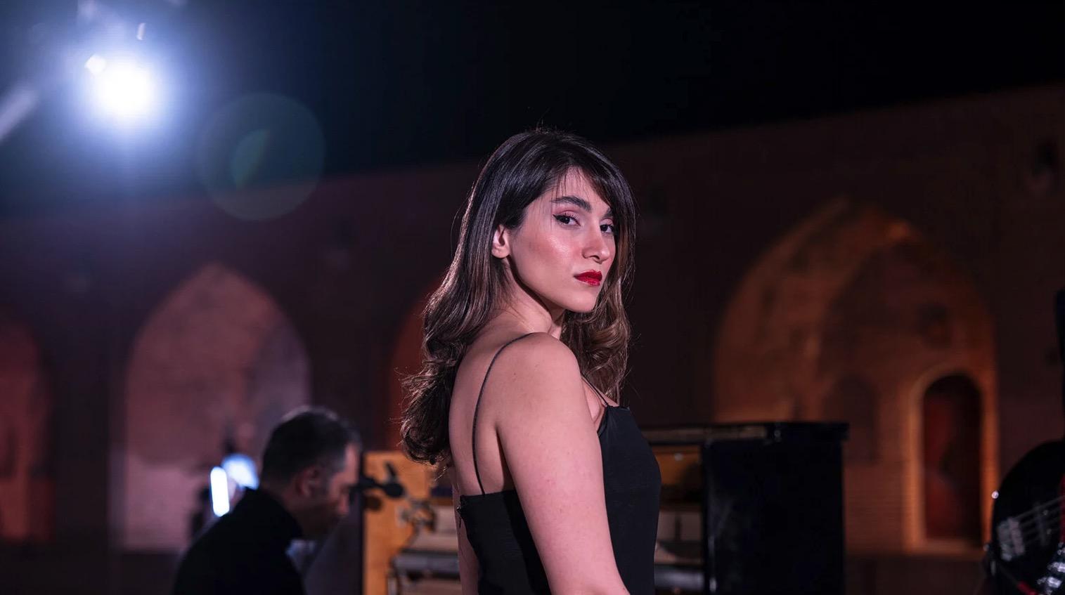 A woman with long hair and red lipstick stands in a black dress, looking over her shoulder at the camera. Behind her, a person plays piano under dramatic lighting, with arched structures in the background.