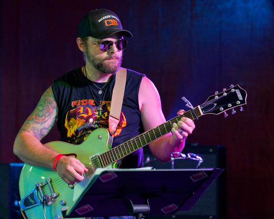 Man with black billed cap, black tank top, and light colored guitar with a little smoke in the background