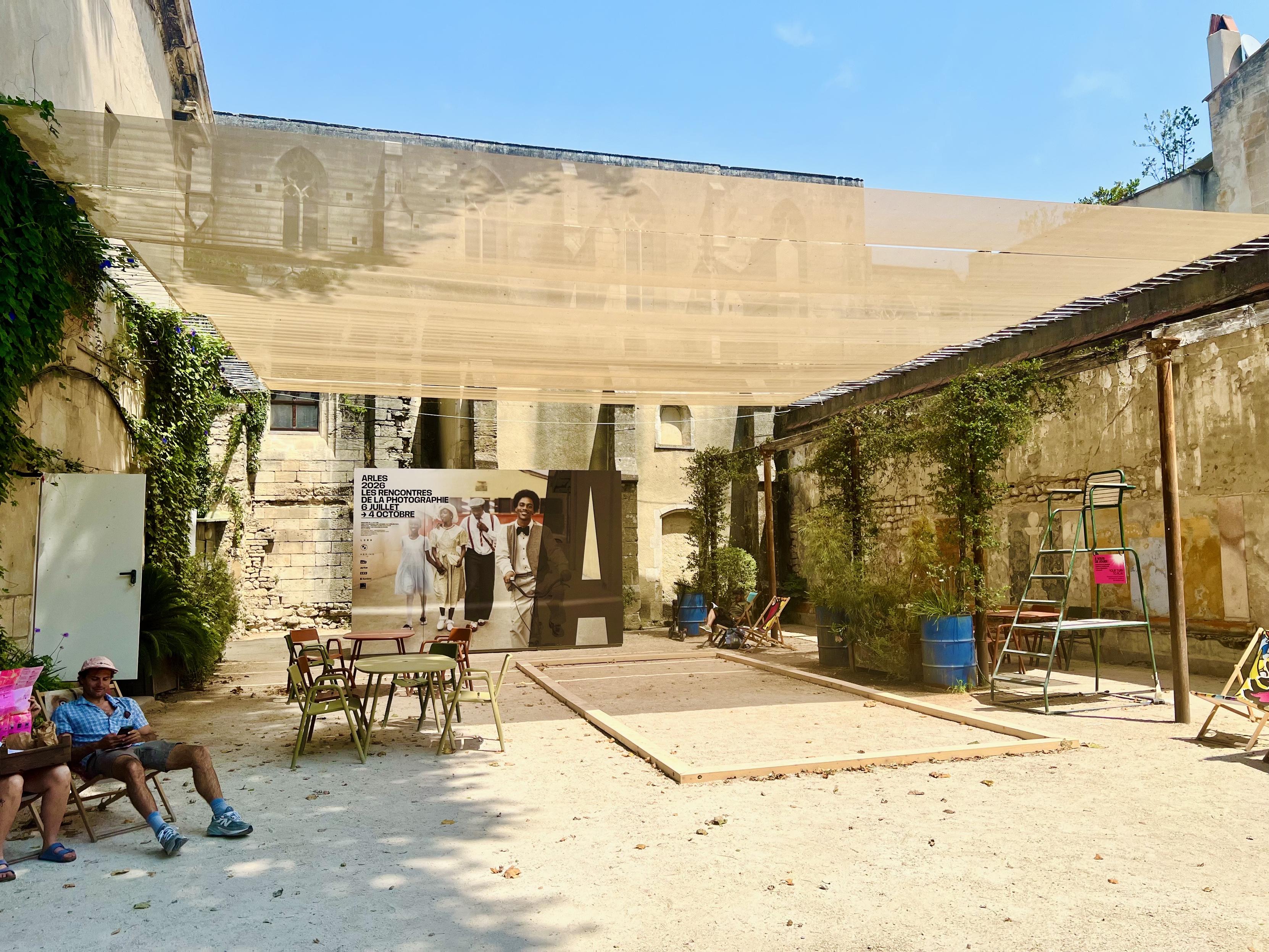 ‎⁨Arles⁩, ⁨Côte Camarguaise⁩, ⁨France⁩

A sunny outdoor courtyard featuring stone walls and a light beige shade sail stretched overhead. In the center, a wooden frame defines a pétanque court on the gravel ground, surrounded by green metal tables and chairs, potted plants, and lounge seating. In the background stands a large photography exhibition poster, while people sit relaxed in the shaded area in the foreground.
