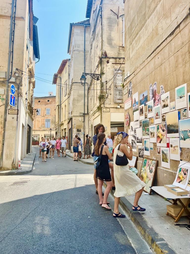‎⁨Arles⁩, ⁨Côte Camarguaise⁩, ⁨France⁩

A narrow, sunlit European street flanked by tall stone buildings, where pedestrians stroll along the pavement. On the right, people stand along the sidewalk examining a outdoor gallery of photographic prints and artwork displayed across the exterior building wall.
