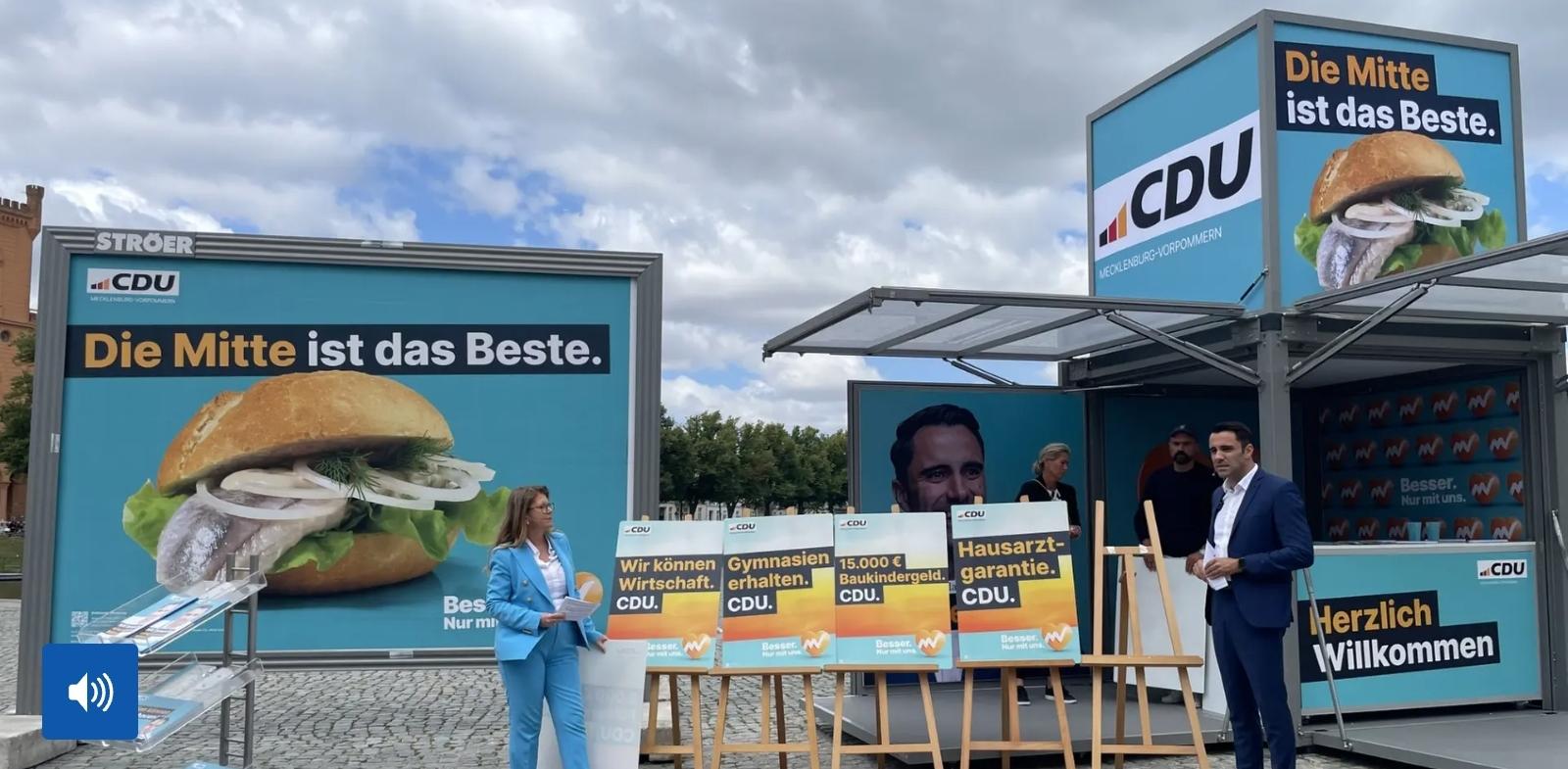 Bildbeschreibung von KI:
Der Screenshot zeigt einen Wahlkampfstand der CDU auf einem gepflasterten Platz unter einem bewölkten Himmel. Links steht eine große Werbetafel mit einem hellblauen Hintergrund. Darauf ist ein Fischbrötchen mit Salat und Zwiebelringen zu sehen. Darüber steht der Slogan Die Mitte ist das Beste. In der linken oberen Ecke befindet sich das Logo der CDU Mecklenburg Vorpommern.

Rechts steht ein moderner Pavillon mit mehreren Werbeflächen im gleichen Design. Auf einer hohen Tafel ist erneut der Slogan Die Mitte ist das Beste neben dem Fischbrötchen zu sehen. Im Inneren des Pavillons hängt ein großes Porträt eines Mannes. An der Seitenwand steht Herzlich Willkommen. Im Hintergrund sind weitere Plakate mit dem Wahlslogan Besser Nur mit uns zu erkennen.

Vor dem Pavillon stehen mehrere Plakatständer mit politischen Forderungen. Zu lesen sind unter anderem Wir können Wirtschaft CDU Gymnasien erhalten CDU 15000 Euro Baukindergeld CDU und Hausarztgarantie CDU.

Im Vordergrund stehen eine Frau in einem hellblauen Hosenanzug mit Unterlagen in der Hand sowie ein Mann in einem dunkelblauen Anzug mit weißem Hemd. Im Hintergrund sind weitere Personen zu sehen. Links unten ist das Symbol eines Lautsprechers eingeblendet, was darauf hinweist, dass der Screenshot aus einem Video stammt.