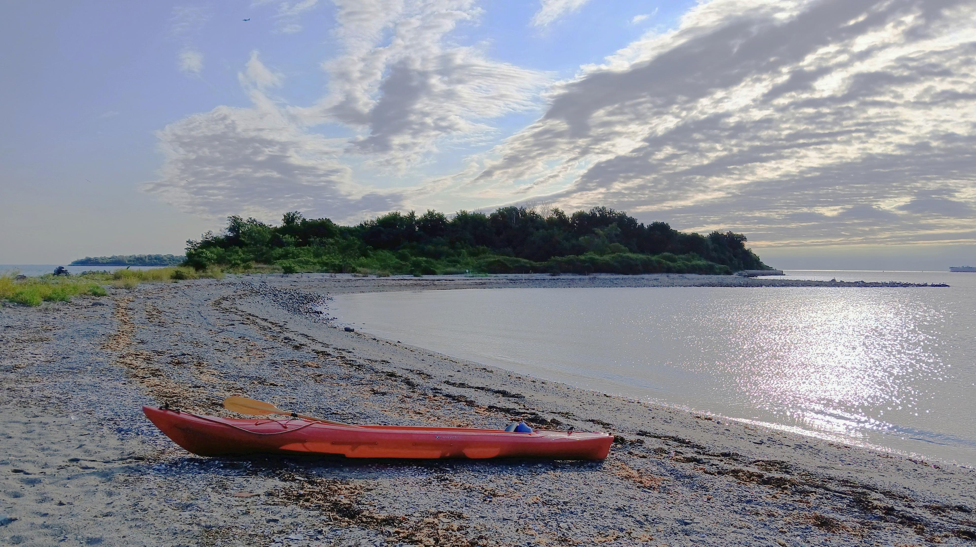 Gravel cove on a sunny/cloudy day, with a red kayak in the foreground and a low wooded hill in the background.