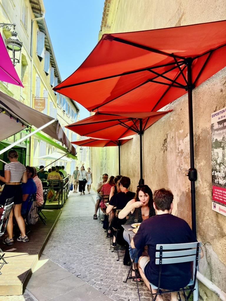 ‎⁨Arles⁩, ⁨Côte Camarguaise⁩, ⁨France⁩

A narrow cobblestone alleyway lined with outdoor restaurant seating on both sides. On the right, diners sit at small bistro tables beneath a row of large red patio umbrellas mounted against a weathered stucco wall, while pedestrians walk down the path between the dining areas under bright daylight.