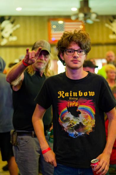 Younger man on right with a black rainbow rising shirt and moderate length hair and behind him and to the left an older man with longer hair making the sign of the horns with an American flag high and near the center
