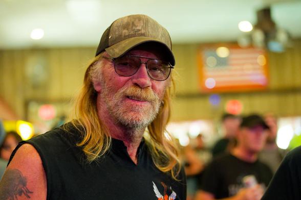 Close shot of man with hat and dark glasses, beared and long blonde hair and tank top